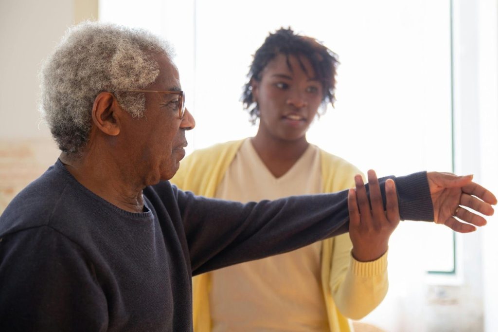 Caregiver for elderly adults helping an older man with gentle arm stretches at home.