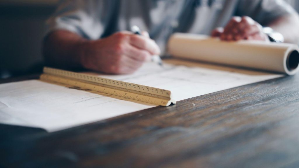 Close-up of an industrial engineer drafting technical specifications for electric motors on a large paper blueprint with a scale ruler, emphasizing accuracy in motor distribution and project planning.